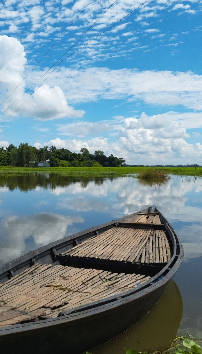 A serene wooden boat floats on a reflective lake in Pabna, Bangladesh, under vibrant clouds and blue sky.