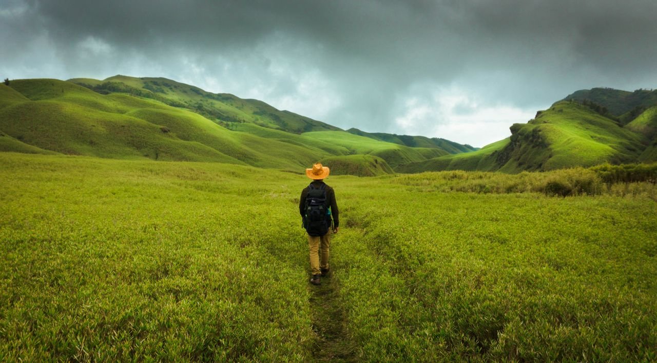 Photograph of a solitary traveler walking through vibrant green hills under a moody sky.